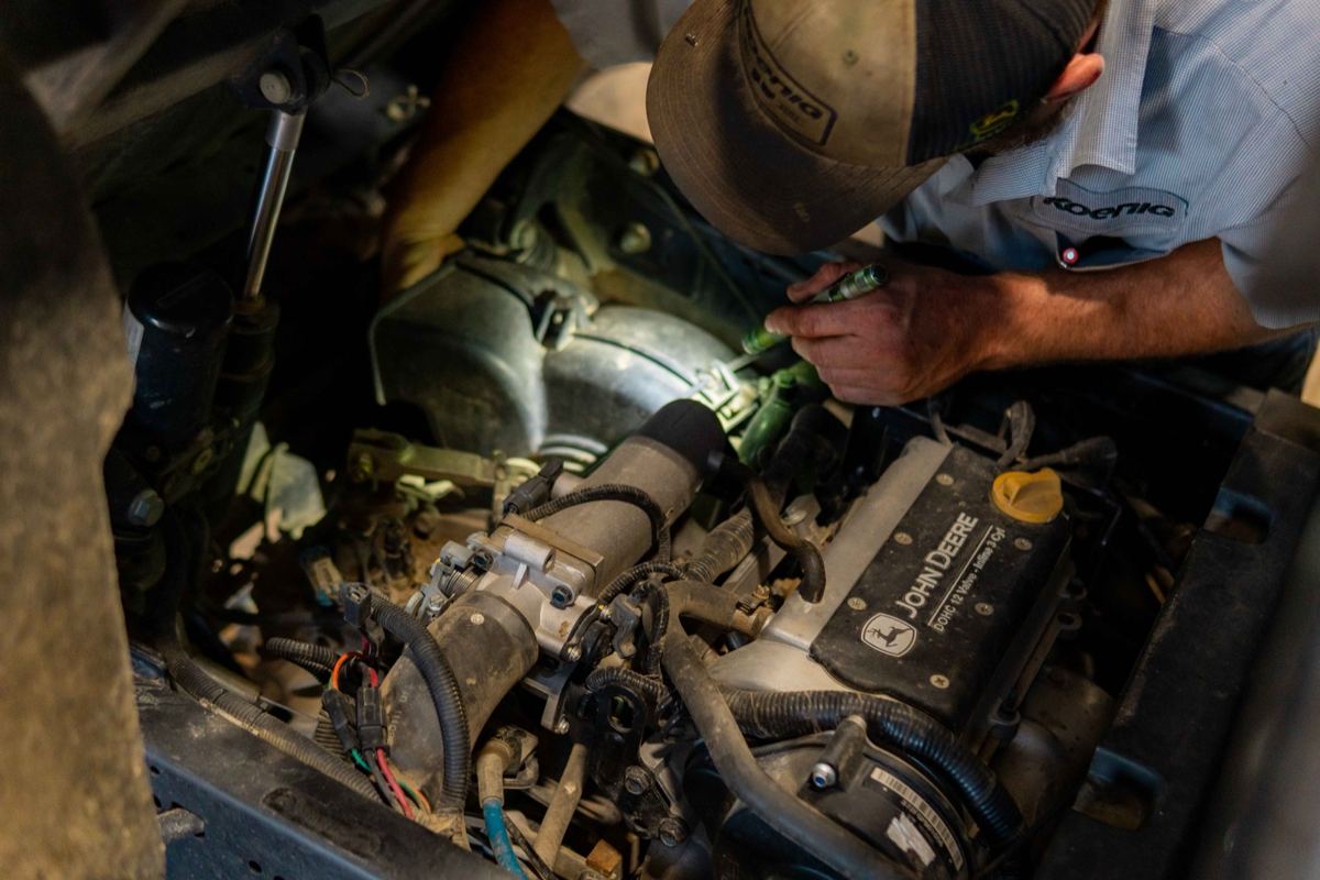 A mechanic in a Koenig shirt inspecting a John Deere DOHC 12 Valve engine with a flashlight