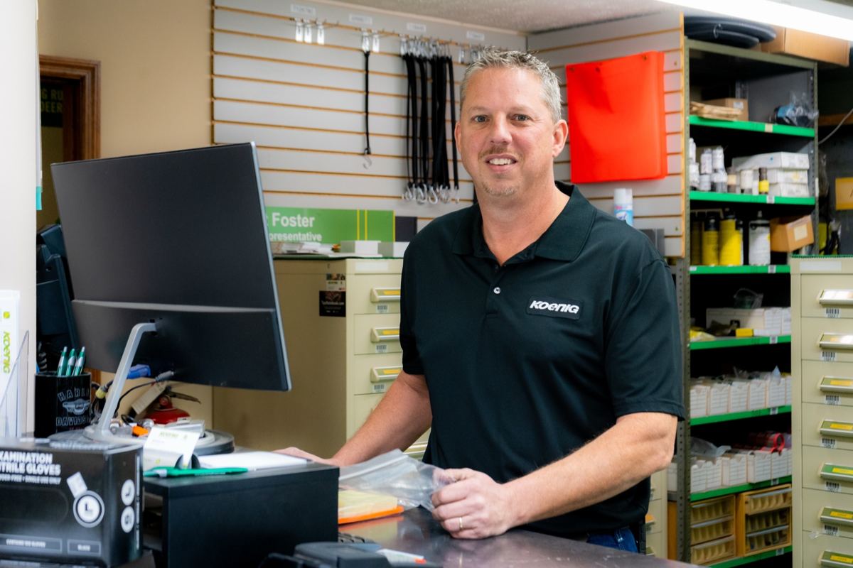 Smiling man in Koenig polo shirt stands behind a counter with a computer and shelves of inventory