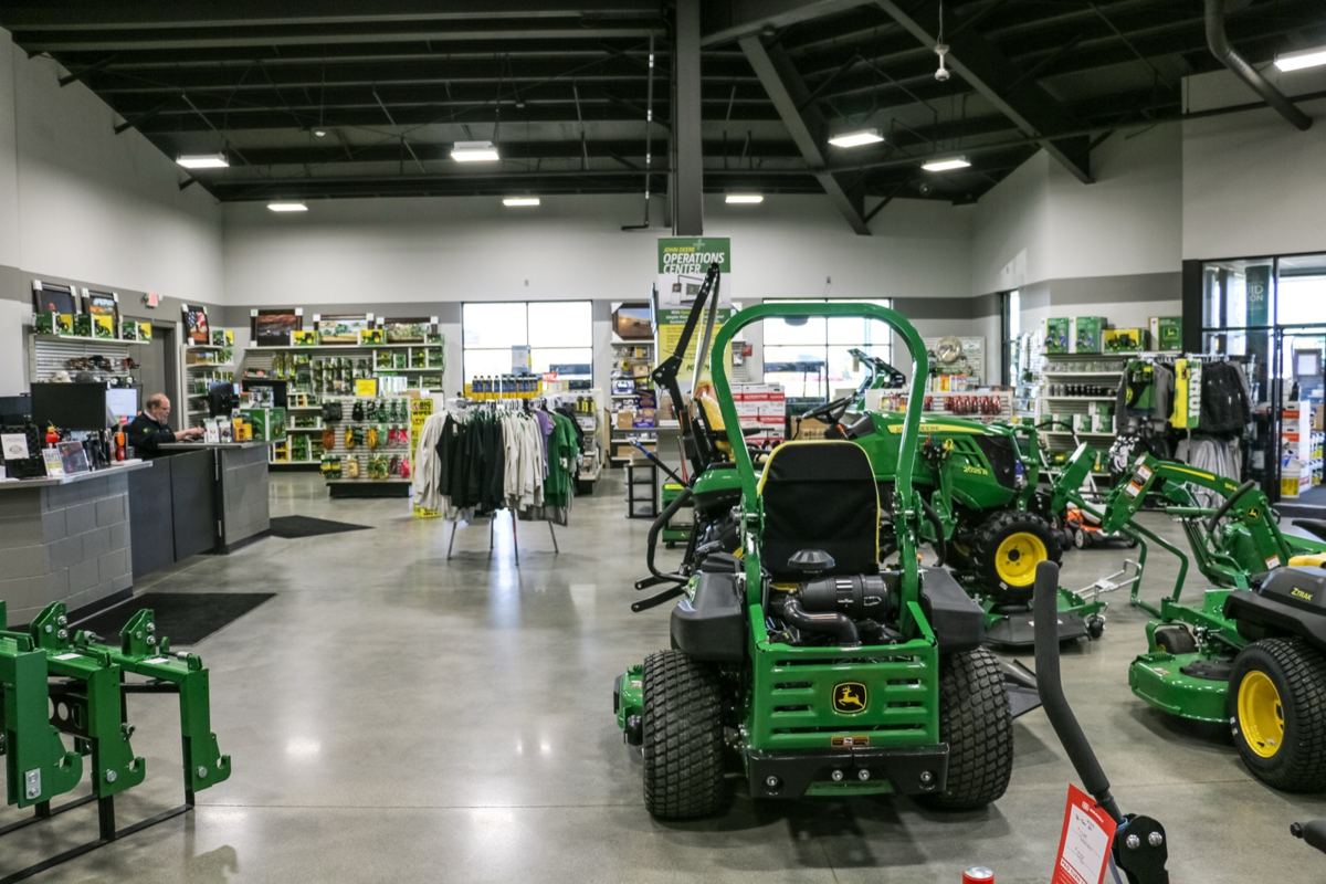 John Deere dealership interior showing green mowers, tractors, parts, clothing, and a man at a service desk