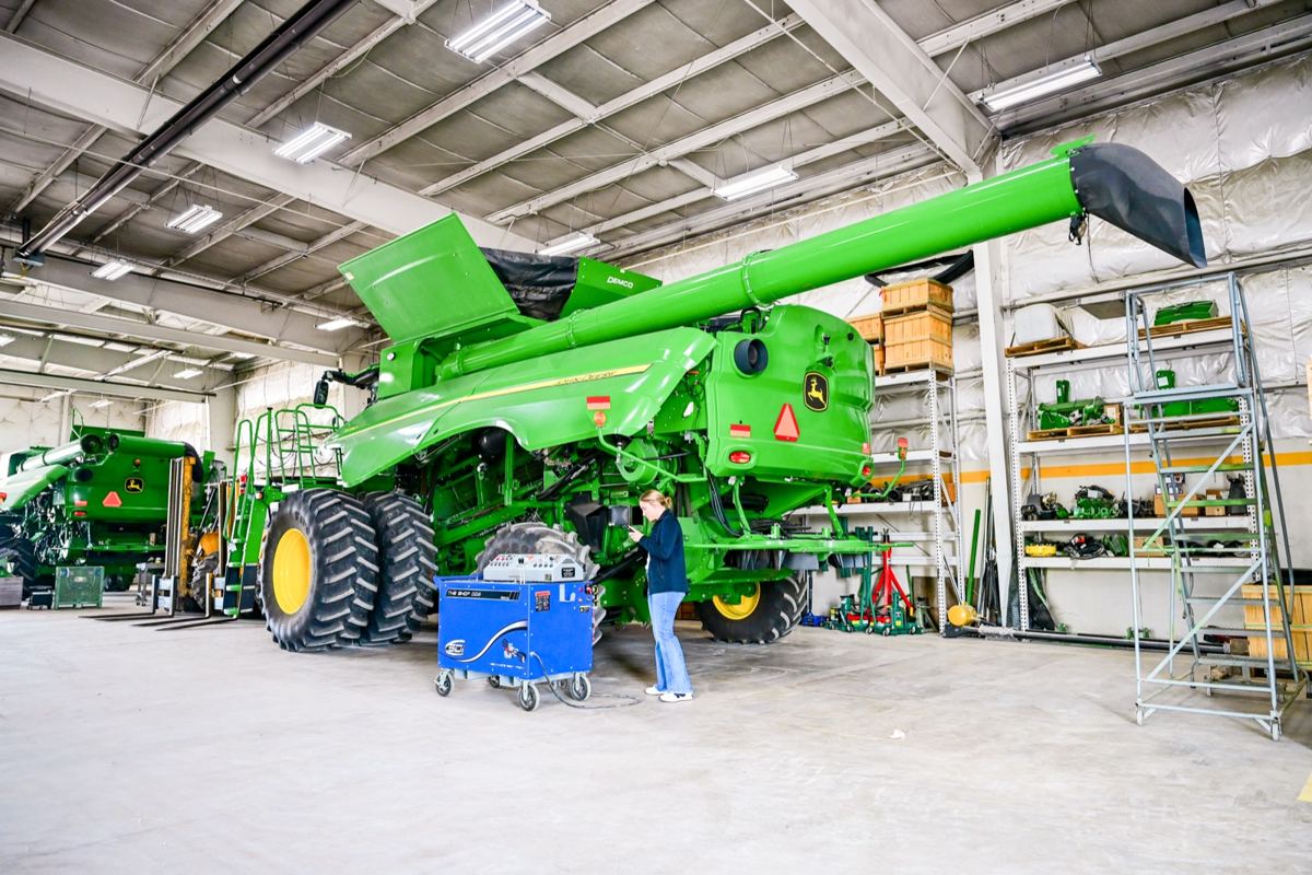 Woman inspecting a John Deere S780 combine harvester in a service bay with another machine and parts shelves