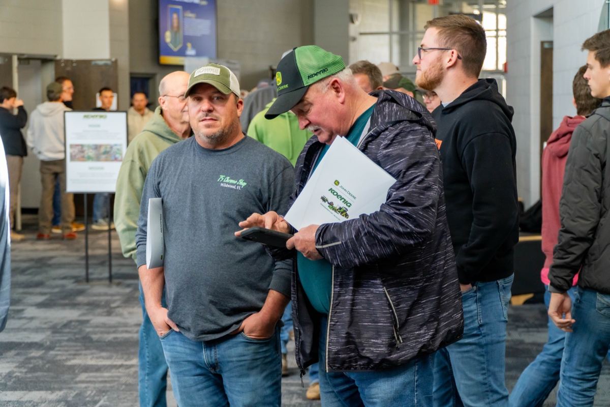 Two men at an indoor event, one in a John Deere Koenig hat holding a brochure and tablet