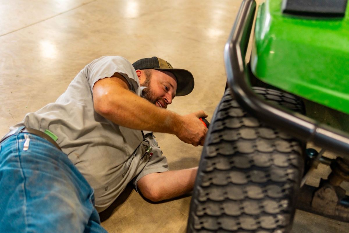Bearded man in a cap and jeans lying on the floor, using a red tool on a green John Deere machine's wheel