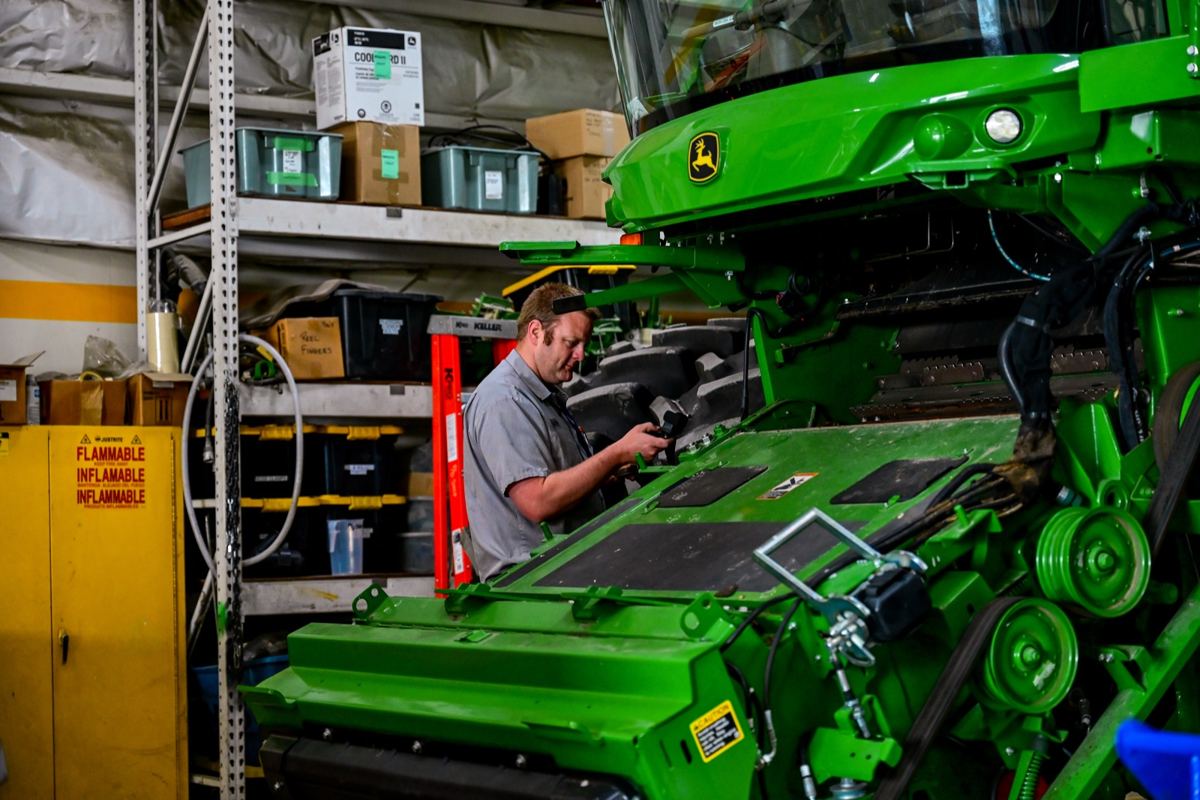 Man working on a green John Deere combine harvester in a service bay with shelves and tools