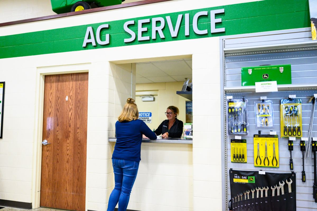 Two women at an AG SERVICE counter with John Deere tools on display and green equipment above