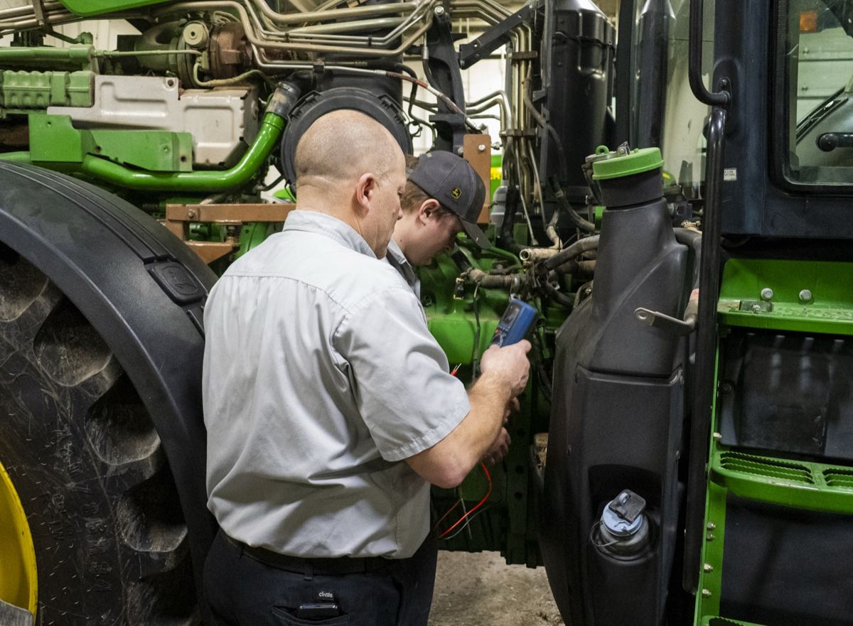 Two technicians using a multimeter to inspect a green John Deere tractor's engine