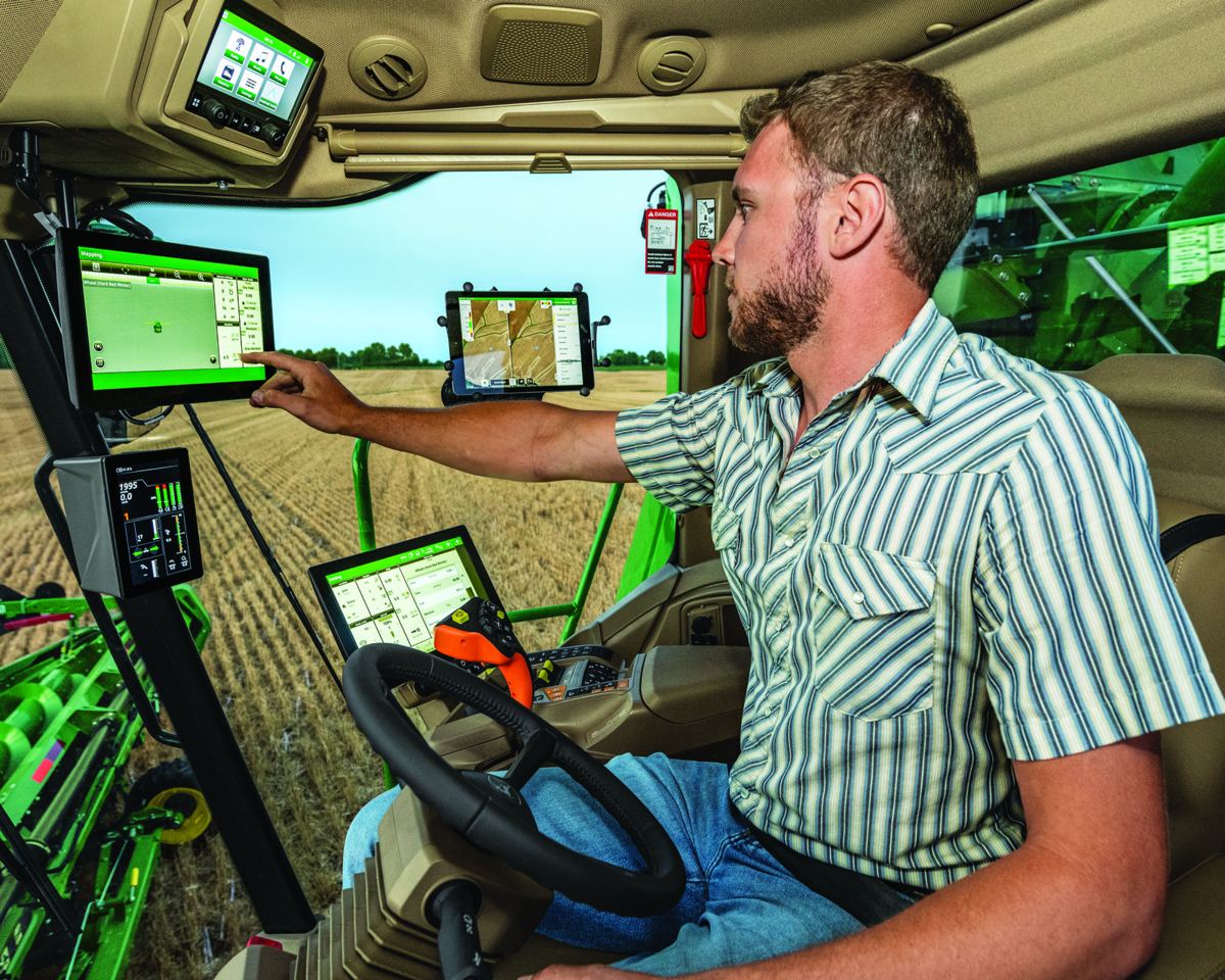 Man in a John Deere combine cab pointing at a green display screen, with other monitors and a field outside