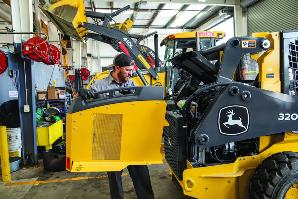 Man servicing a John Deere 320 skid steer, holding its yellow side panel, with a John Deere 410L in the background