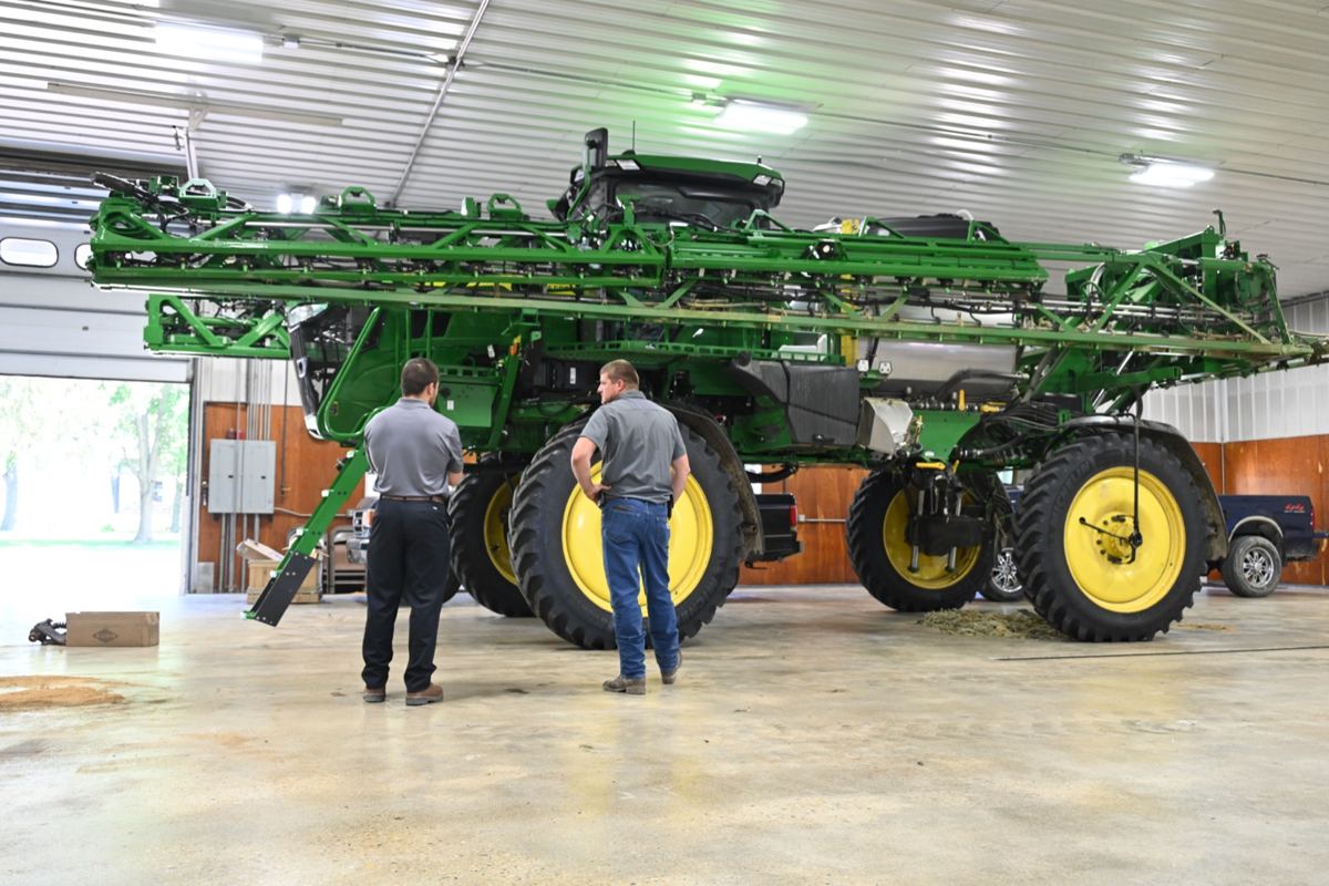 Two men inspecting a large green and yellow John Deere agricultural sprayer with extended booms in a workshop