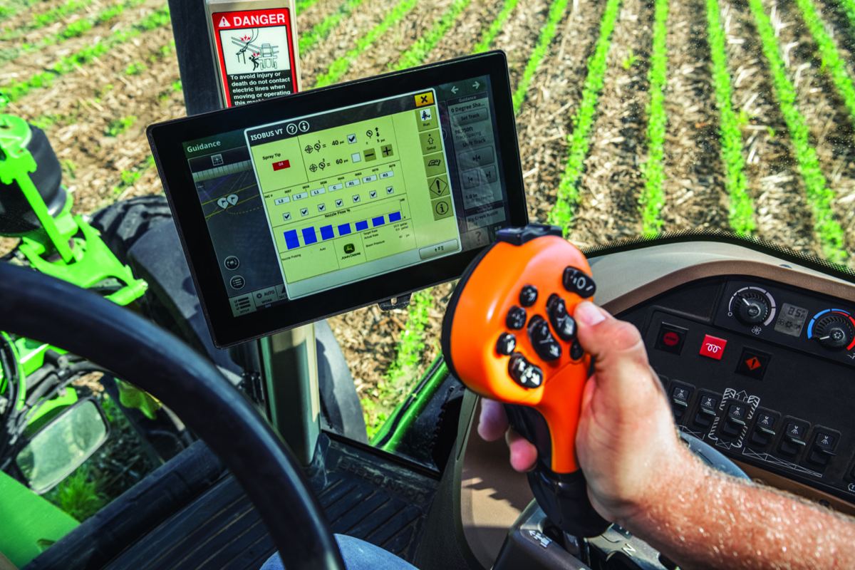 Person's hand operating an orange joystick controller in a tractor cab with a digital display and green crops in the field