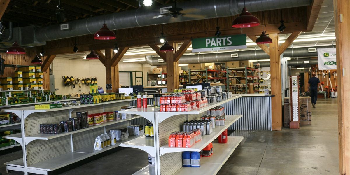 John Deere dealership interior with aisles of parts, fluids, and toy tractors, a 'PARTS' sign, and a man walking