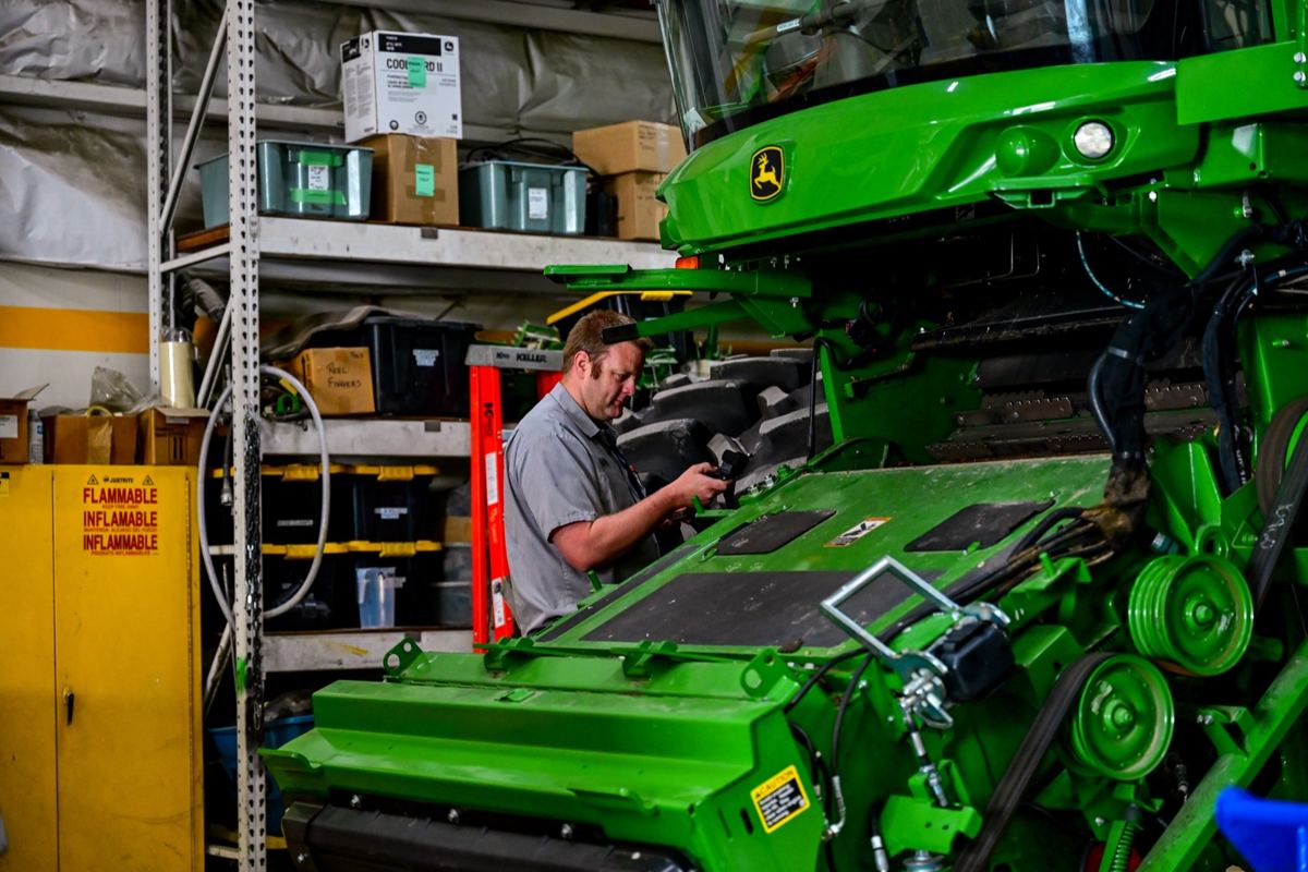 Man in grey shirt with headlamp working on a large green John Deere agricultural machine in a service bay
