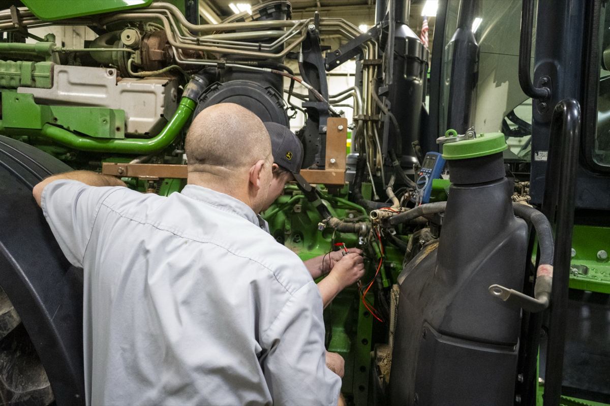 Two technicians are working on the engine of a large green tractor in a service bay, performing diagnostics or repairs with wires connected to components