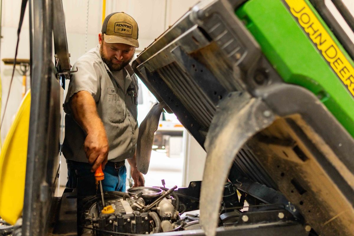 Bearded man in Koenig hat using an orange screwdriver to repair a John Deere vehicle engine