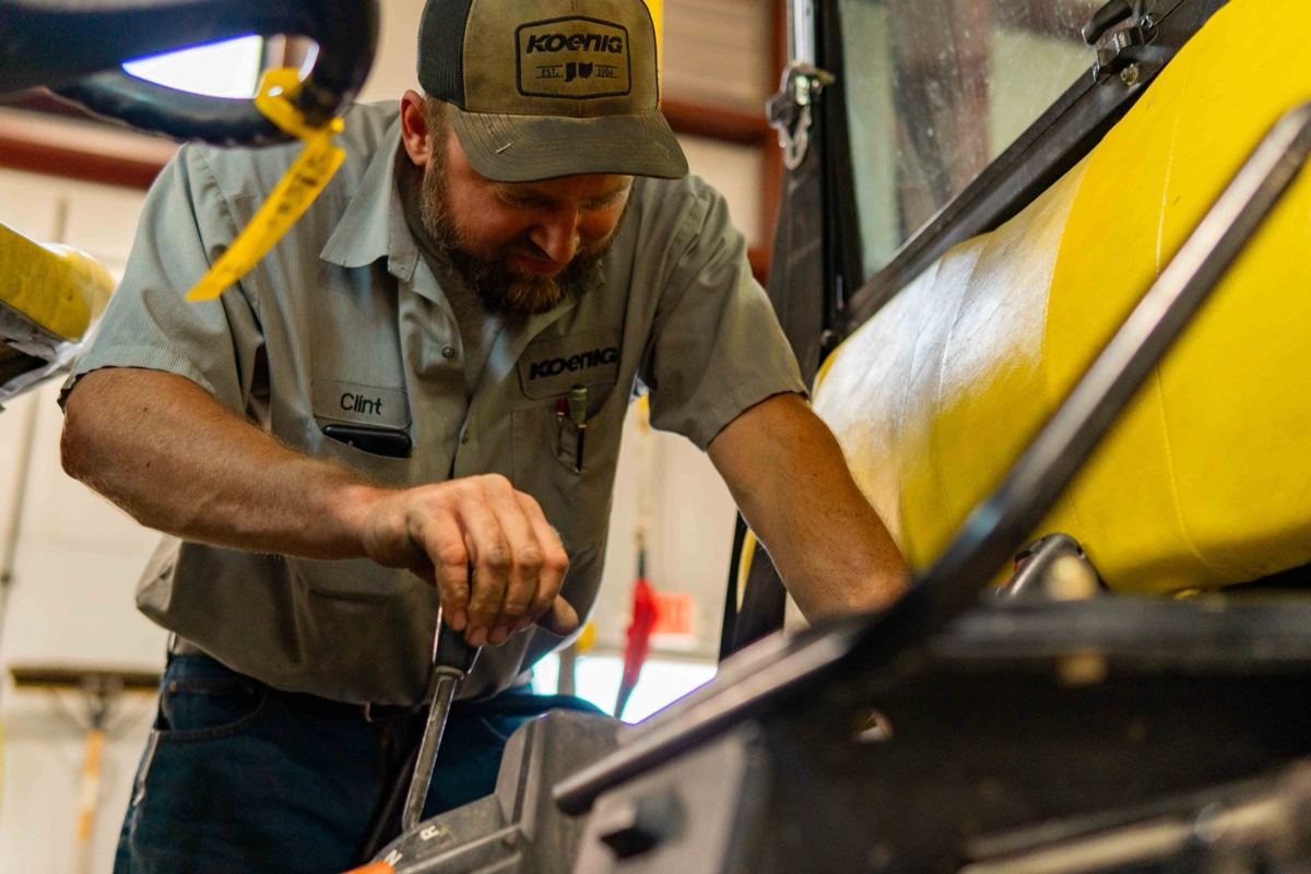 A Koenig Equipment technician is working on a component of heavy machinery in a service shop, implying inspection or repair, though the specific component is not identified as tillage blades or rolling baskets