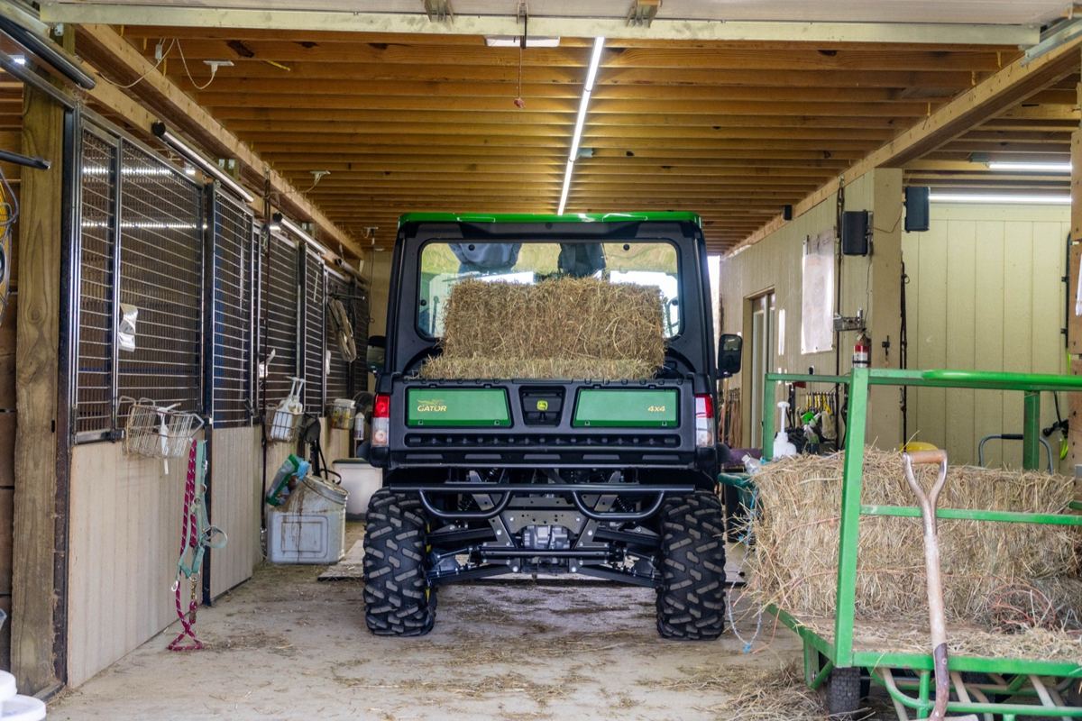 John Deere Gator 4x4 utility vehicle with a bale of hay in its bed, parked in a barn aisle with horse stalls and a hay cart