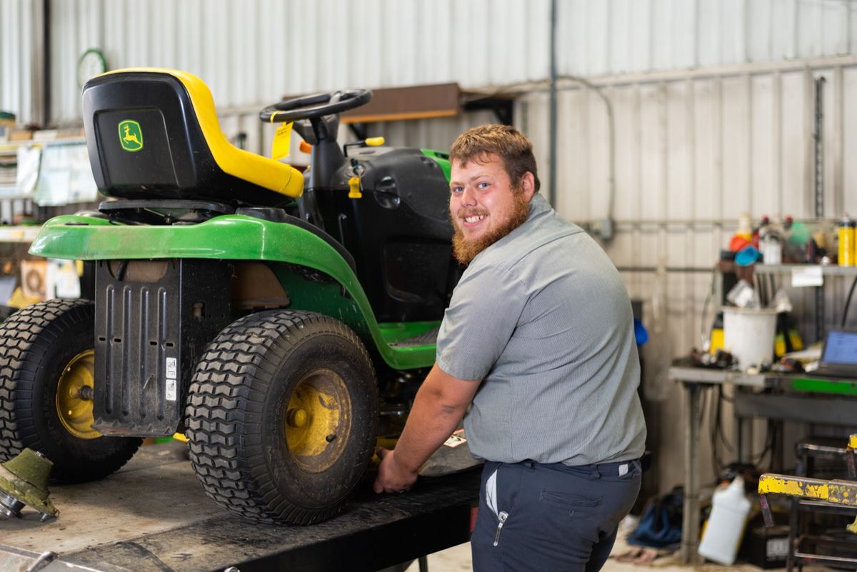 Smiling man in a grey shirt working on a green and yellow John Deere riding lawn mower in a workshop