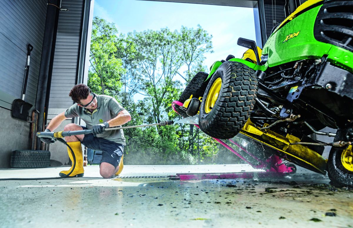 Man pressure washing the underside of a green and yellow John Deere X304 lawn tractor