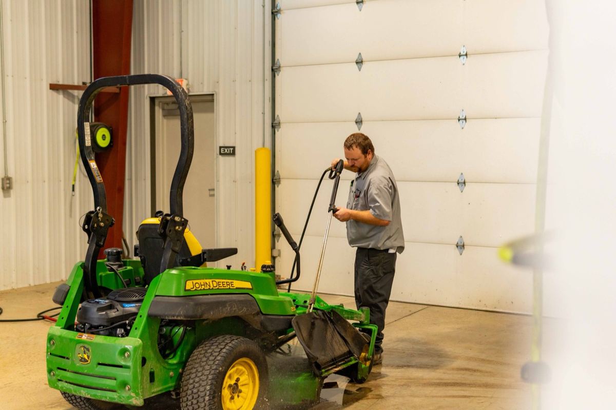 Man in a gray shirt pressure washing a green John Deere zero-turn mower in a service bay