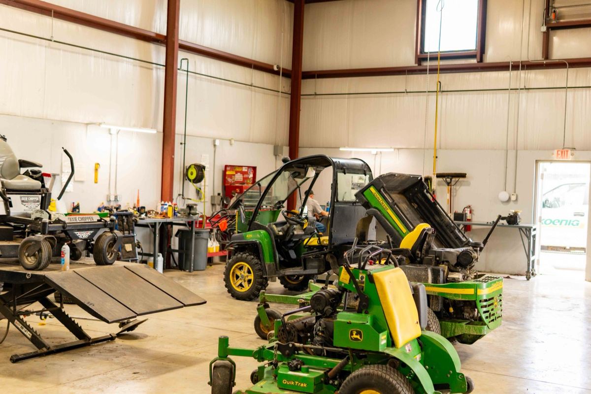 John Deere Gators, a QuikTrak mower, and a zero-turn mower on a lift in a service bay