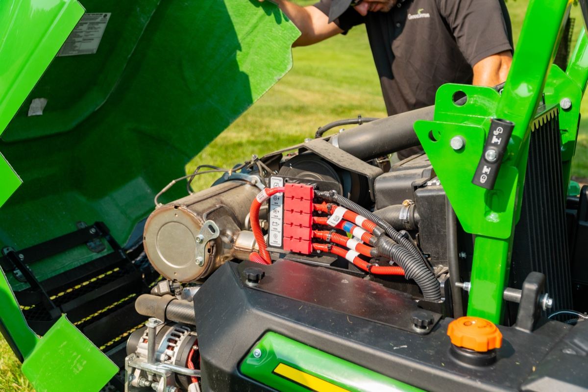 Man inspecting the engine of a green John Deere machine with an open hood, revealing a red fuse box and wiring