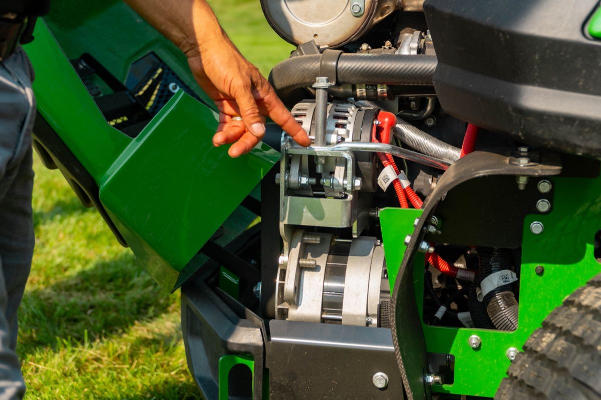 A person's hand points to a silver engine component with a black belt and red wires on green and black machinery