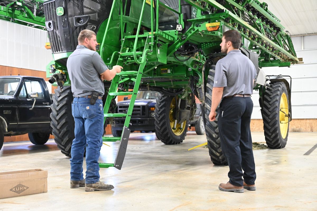 Two men discussing next to a large green John Deere self-propelled sprayer in a workshop