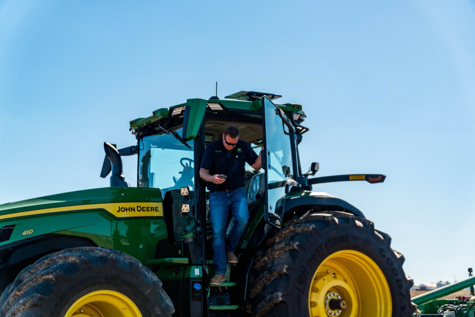 A farmer steps down from a John Deere tractor while checking his phone