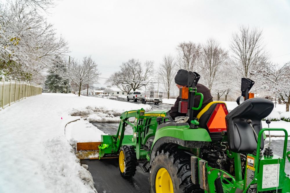 A John Deere compact tractor with a front blade clearing a snowy drive