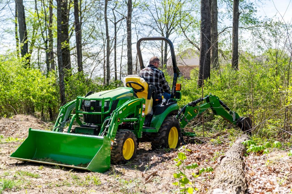 A John Deere 2025R with loader and backhoe clearing brush in a wooded lot