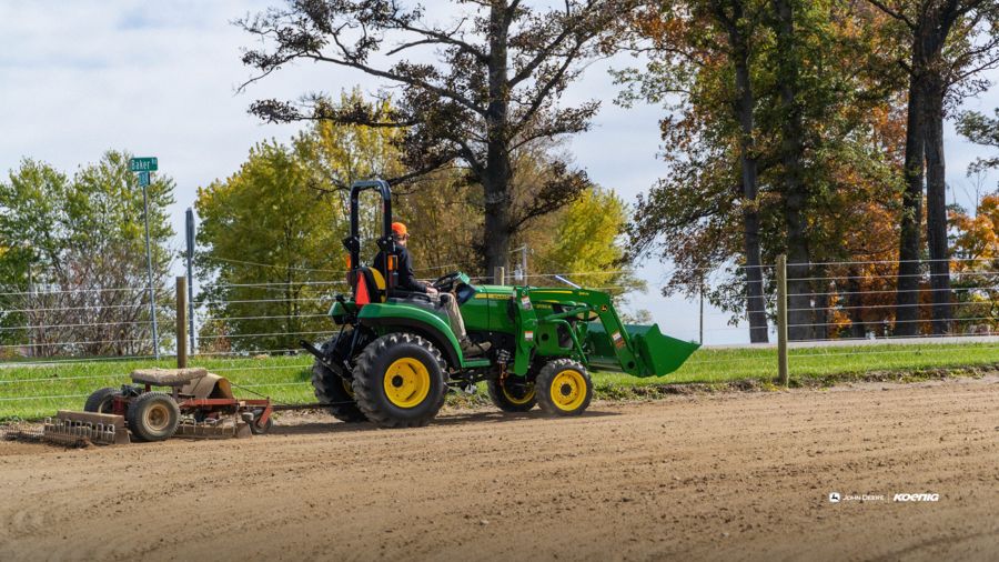John Deere compact utility tractor with a front loader pulling an arena groomer past autumn trees, desktop wallpaper preview