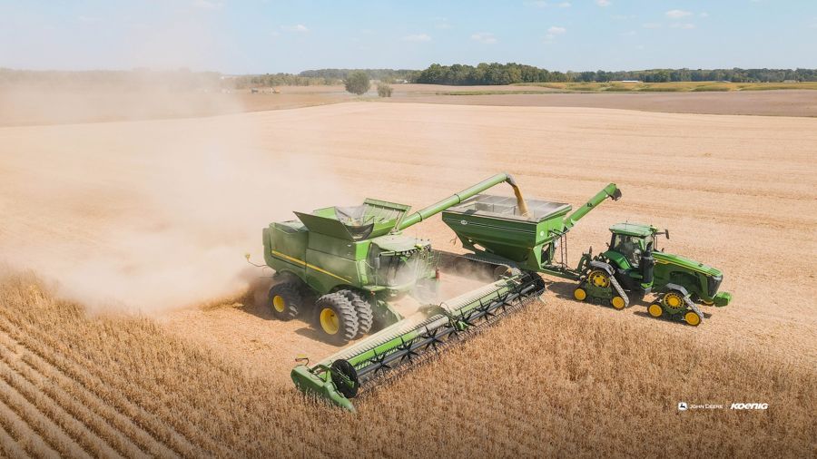 An aerial view shows a combine harvester unloading grain into a grain cart pulled by a tractor in a field under a blue sky., desktop wallpaper preview
