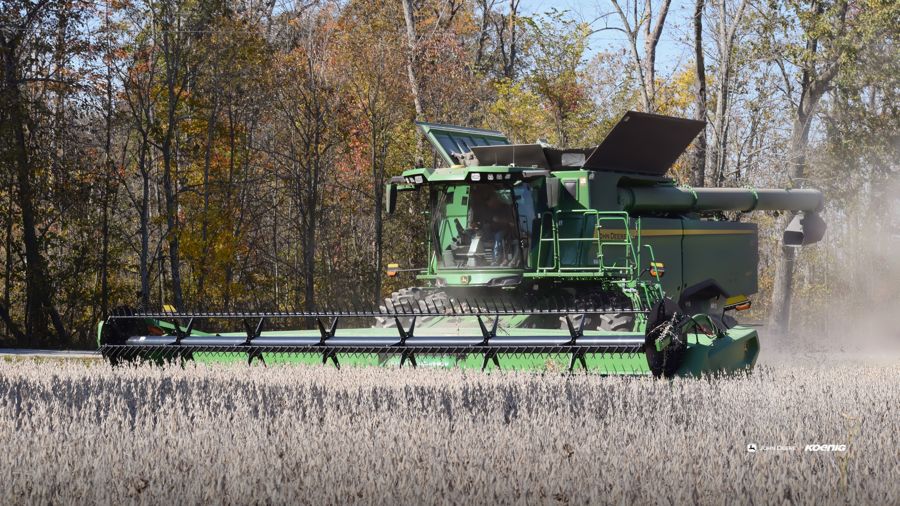A green John Deere combine harvester is actively harvesting crops in a field under a clear sky, with a line of autumn trees in the background., desktop wallpaper preview