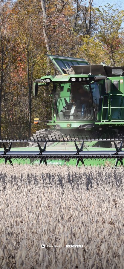 A green John Deere combine harvester is actively harvesting crops in a field under a clear sky, with a line of autumn trees in the background., phone wallpaper preview